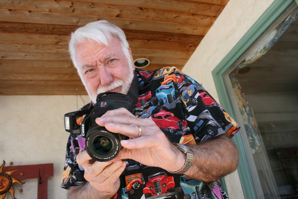 A photo of John Schulz wearing a shirt with colorful cars, holding a camera with the lens facing forward.