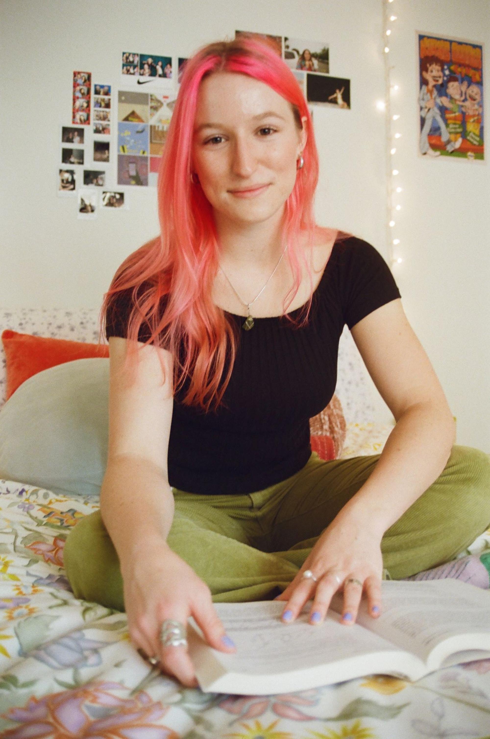 Grace, the person behind ChureThing, is sitting on a bed reading a book. She has pink hair and is wearing casual clothing.